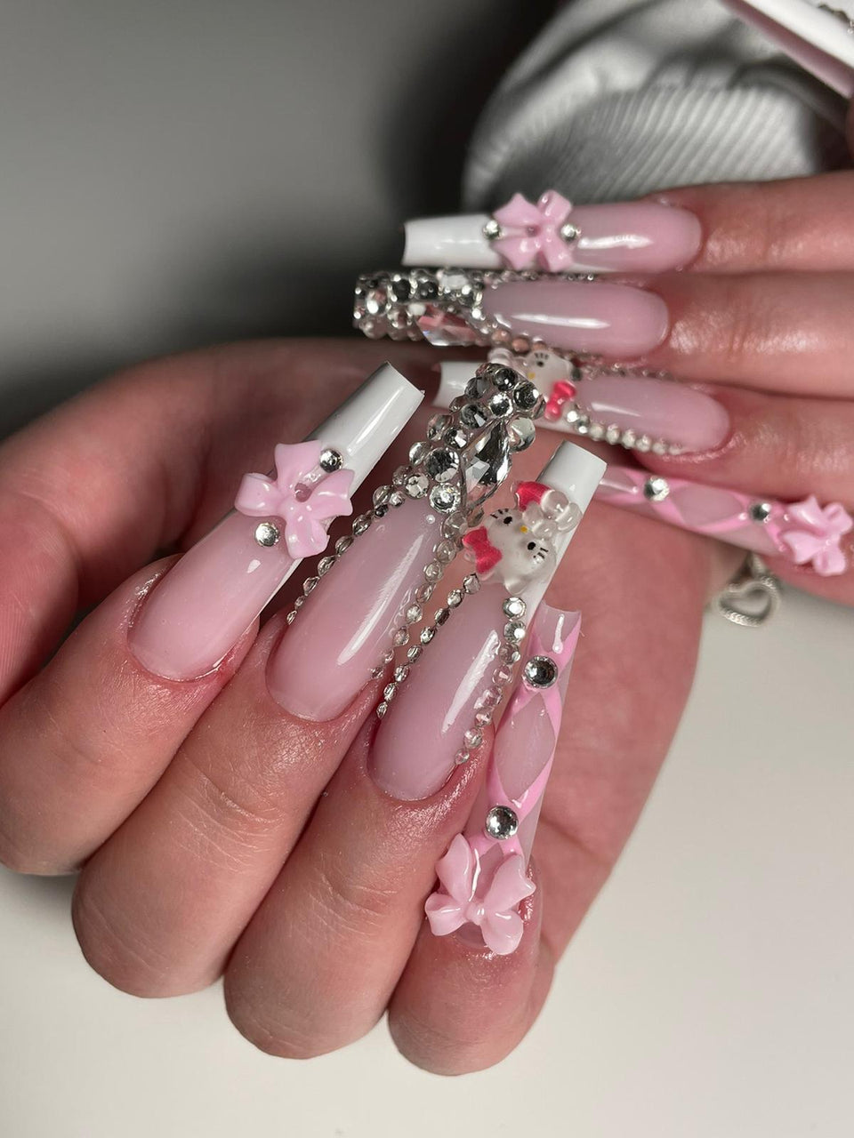 Nail art rose et blanc sur ongles longs, avec strass, nœuds et décorations en relief.