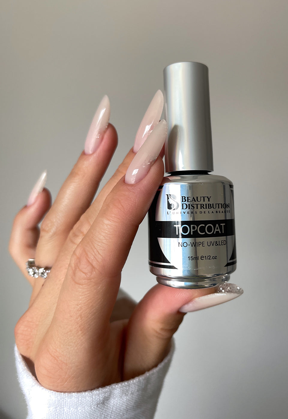 Main aux ongles longs nude tenant le Top Coat No-Wipe UV/LED Beauty Distribution 15 ml.